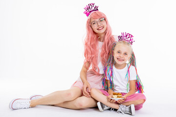 Delighted preschooler daughter with colorful braids sitting in lotos pose wearing plain t-shirt and pink skirt posing with modern young mother with pink wig and glasses over plain white background.