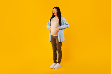 Fototapeta na wymiar Full body length shot of pregnant woman suffering from abdomen pain, touching belly and breathing, yellow background