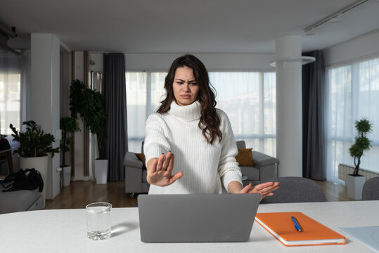 Young shocked businesswoman annoyed unpleasant spam message pop-up ad frantically looking at laptop screen while sitting and working from home trying to cover monitor with hands, inappropriate content