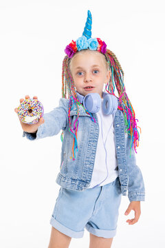 Adorable little girl with colorful barids and unicorn headband with flowers and blue corn standing on white background wearing denim clothes and stylish blue headphones showing donut in hand.