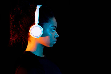 Young black lady in wireless headphones, listening to music with in neon light, side view, banner design with free space