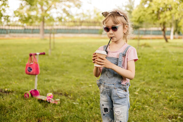 Fototapeta na wymiar little girl drinking cocoa in the summer in the park wearing a cat ear headband and sunglasses