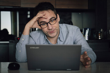 Young male in casual clothes concentrated guy adjusting eyeglasses while working on laptop and looking at pc screen during remote work from home office, overworking man feeling tired, closeup