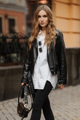Fototapeta na wymiar Fashionable blonde woman model with black leather jacket and style bag walking the city street