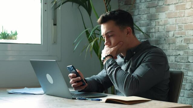 Confident man sitting at desk taking break in work with electronic documents on laptop to make answer telephone call. Smiling young guy freelancer synchronize data between home computer and smartphone