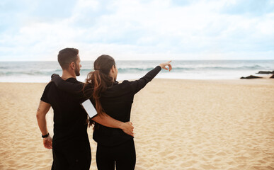 Young sporty couple working out together at the beach by seaside, resting after training, embracing, woman pointing away