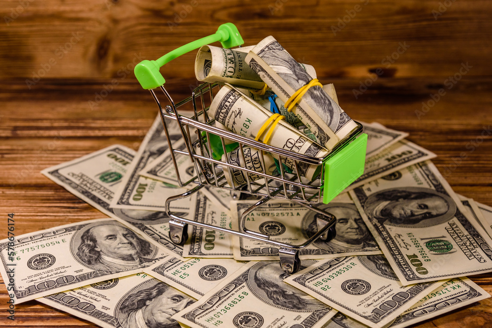 Wall mural Small shopping cart with rolled up one hundred dollar banknotes on a wooden background