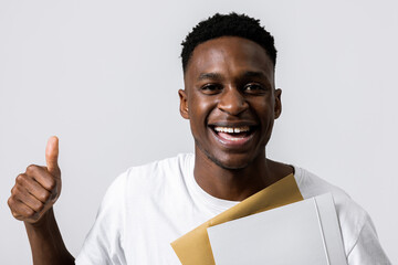 Close up shot portrait of cheerful handsome african american black man guy holding folder in hands showing thumb up at camera smiling isolated on grey background in studio.