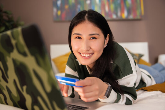 Portrait photo of delighted asian kazachstan girl in casual clothes outfit smiling at camera while laying on bed in front of laptop computer doing purchase shopping online holdin credit card in hands.