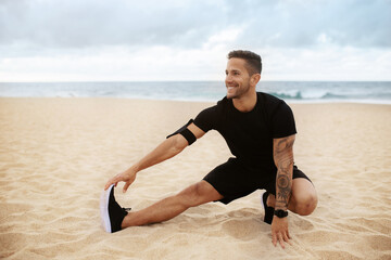 Young strong sporty man in sports clothes doing warm up training, stretching legs on beach by seaside outdoors