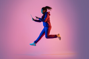 Fototapeta na wymiar Full length of excited black woman with digital tablet jumping up in neon light, studio background, copy space