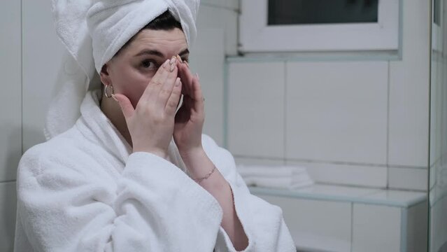 A woman in a bathrobe looks in the mirror and gives herself a facial massage. Touch the skin with your fingertips, enjoy an easy effective anti-wrinkle treatment at home. Removal and prevention of mim