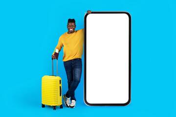 Naklejka premium Travel App. Smiling Black Man With Suitcase Standing Near Big Blank Smartphone