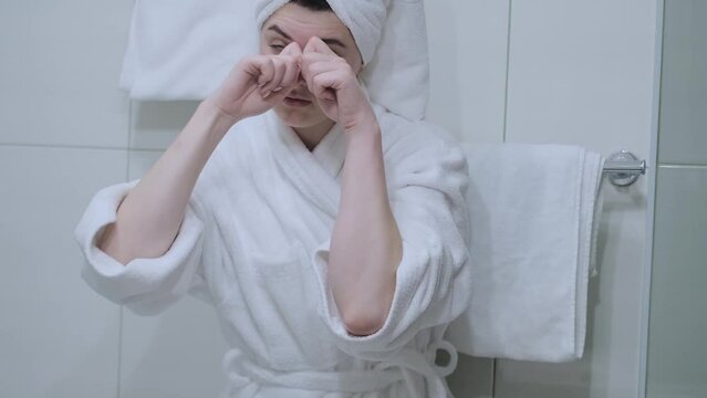A woman in a bathrobe looks in the mirror and gives herself a facial massage. Touch the skin with your fingertips, enjoy an easy effective anti-wrinkle treatment at home. Removal and prevention of mim