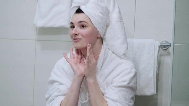 A woman in a bathrobe looks in the mirror and gives herself a facial massage. Touch the skin with your fingertips, enjoy an easy effective anti-wrinkle treatment at home. Removal and prevention of mim