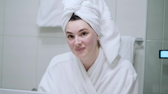 Close-up of Woman with Smooth Healthy Skin, Looking in Mirror, Touching her Face Gently, Smiling. Portrait of Girl Model in White Bathrobe and Towel Posing for Camera. Skincare Concept.