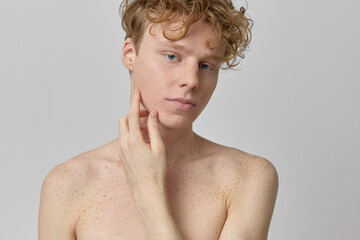 Beauty portrait of shirtless handsome curly and red haired male model with freckles touching his face with hand looking at camera with strong face and clear skin over grey background  in studio shot.