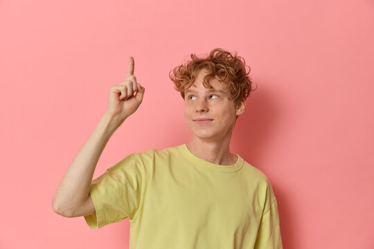 Studio waist-up shot of a cute red haired freckled teenager smiling looking upwards pointing his finger up comes up with the idea has a brainwave shows gesture on pink background for your advertising.