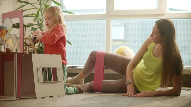 Mother does sport exercises while daughter plays with dolls toy house, pretty blond kid, women wearing bright clothes. Trying to find time for sports being a parent, using sport band. 4k footage
