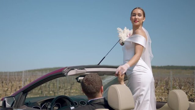 A young bride looks out of the car. Action. A model in a white dress with red lips holds a wedding bouquet in her hands and poses against a blue sky.