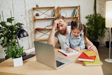 Mature man, father sitting at table and working on laptop remotely at home with his little preschool daughter drawing with him. Concept of fatherhood, childhood, family, freelance job, home office