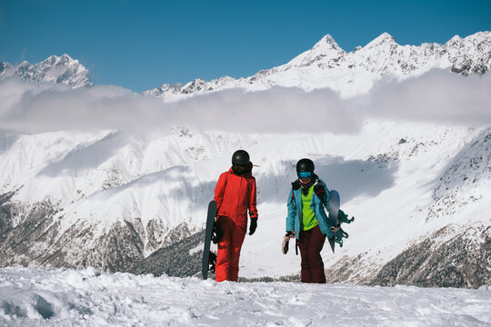 Couple of friends snowboarders walking on mountain slop with snowboards, mountains peaks covered snow on background
