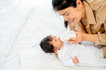 Young Asian mother give milk in bottle to newborn baby lie on bed and take care with happiness in bedroom of their house.