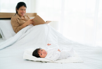 Newborn baby lie on bed with relax and happiness while her Asian mother sit and read book in bedroom in background.