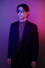 Fototapeta premium Portrait of confident young man in suit and sunglasses standing against pink and purple background