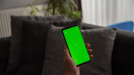 Back view of young europen woman who is using chroma key smartphone screen.She is sitting on the sofa in her cozy living room.
