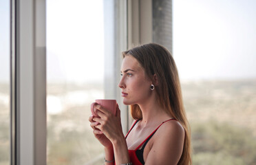 young woman drinks from a cup and looks at the view from the window