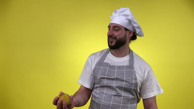 Smiling Bearded Male Chef Preparing For Cooking, Throw Up Apple, Bite Fruit Isolated On Yellow Background. Cheerful Middle-aged Cook Man Eating Fresh Apple Smiling Looking At Camera. Culinary Concepts