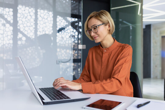 Beautiful Young Businesswoman Inside Office Working With Laptop, Blonde With Short Hair Typing On Keyboard And Smiling, Female Programmer Coding Software While Sitting At Workplace.