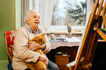 Portrait of an old gray-haired woman artist hugging a cat and sitting near an easel near the window.