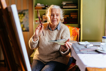 Focused happy old senior retired woman in sweater drawing on easel with paintbrushes holding...