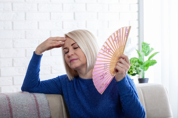 Mature woman experiencing hot flush from menopause. This photo captures the discomfort of hot flashes during menopause, as a woman struggles to cool herself with a delicate paper fan.