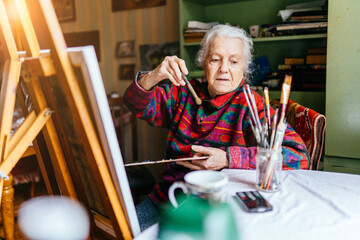 Artist gray-haired senior female drawing sitting on chair at home. Learning new hobbies concept.