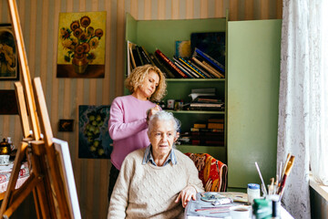 Gray-haired old woman with her caregiver daughter brushing her hair. Senior Caucasian woman painting at easel.
