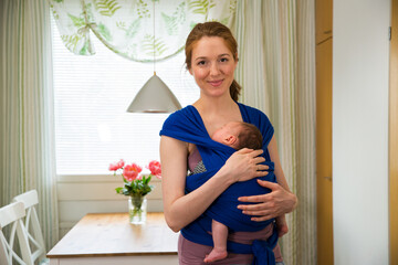 Portrait of a simple woman with baby in a wrap carrier cooking in the kitchen. Interior of a cozy modern kitchen. Real mother doing her chores. 