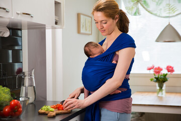 Woman with baby in a wrap carrier cooking in the kitchen, cutting vegetables with knife on a...