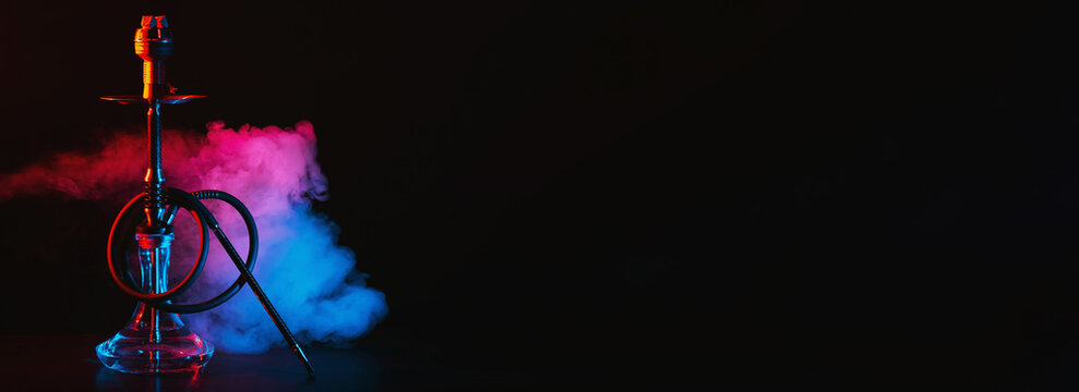 hookah with a glass flask and a metal bowl shisha with colored smoke on the table on a black background with a copy space