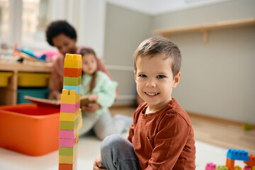 Happy kid stacking toy blocks while playing at kindergarten and looking at camera.