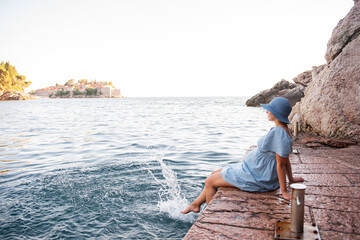 Pregnant woman traveler in denim dress sits by sea, Looks at island of Sveti Stefan in Montenegro