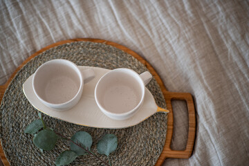 On the Linen bedspread lies a wooden round tray, on it are two empty ceramic cups, green eucalyptus