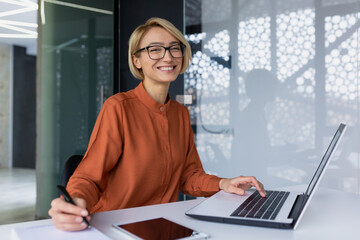 Successful and happy business woman looking at camera sitting inside office working with documents, portrait of satisfied woman writing in notebook and working using laptop at work.
