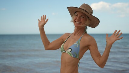 Portrait of smiling happy woman with arms outstretched arms wearing bikini and hat with the blue ocean behind her.
