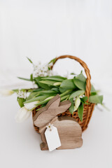 Wooden bunny, pear flowers, white tulips in a wicker basket on a light background.  Easter spring background with bunny and white flowers in light room