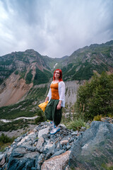 Naklejka premium Happy girl, tourist posing on the top of the mountain, in the background the top of Mount Ushba, Georgia, Svaneti. Travel and vacation concept. Hiking track to the Chalaadi glacier. Vertical photo