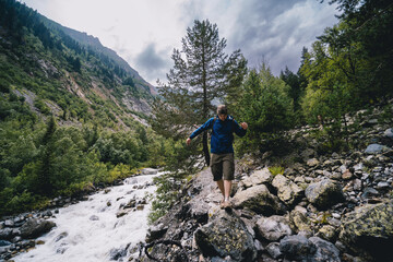 Obraz premium A hiker with backpack, a traveler, walks alone, balances stepping from boulder to stone. Caucasus mountains, The turbulent flow of the river. Svaneti, Georgia. Summer cloud rain season.