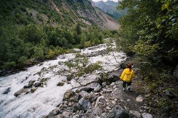 Woman in yellow jacket, tourist hiking track to the Chalaadi glacier. in the background turbulent flow of the river, Georgia, Mestia. Travel and vacation concept.